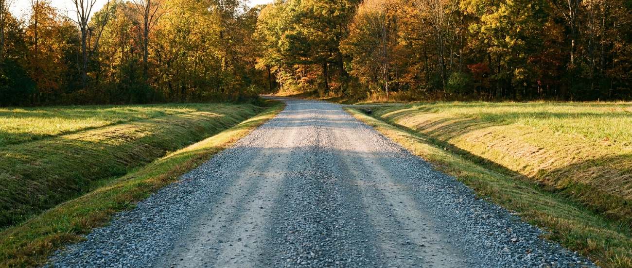 Long crowned gravel driveway running between fields toward a treeline